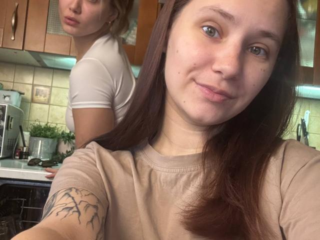 HotCaramelsTrio with brown hair, blue eyes, and a tree tattoo on her arm, wearing a beige shirt, standing in a kitchen, with another HotCaramelsTrio in a white shirt.