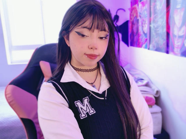 Arieyaara with long brown hair, black choker, white shirt, black vest with "M", eyeliner, and red lipstick, sitting in pink chair, looking down, in purple-lit room.