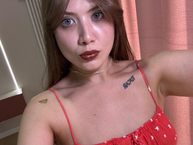 HotCaramelsTrio with shiny skin, red dress, small tits, tattoo "XOXO" on right chest, small tattoo on left shoulder, silver necklace, and pink curtains in the background.
