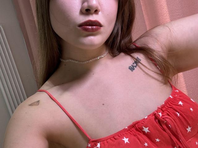 HotCaramelsTrio's face, showing glossy red lips, a silver choker, and a tattoo near the right shoulder, wearing a red star-patterned dress with spaghetti straps, exposing her tits.