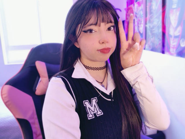 Arieyaara with long brown hair, wearing a black and white school uniform, black choker, and red lipstick, making a peace sign, with a pink and black chair behind.
