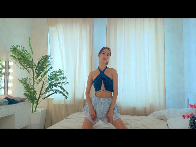 AzucenaMervis with blue halter top showing her tits, and grey lace shorts, kneeling on a bed, hands on her ass, in a bright, softly lit room.