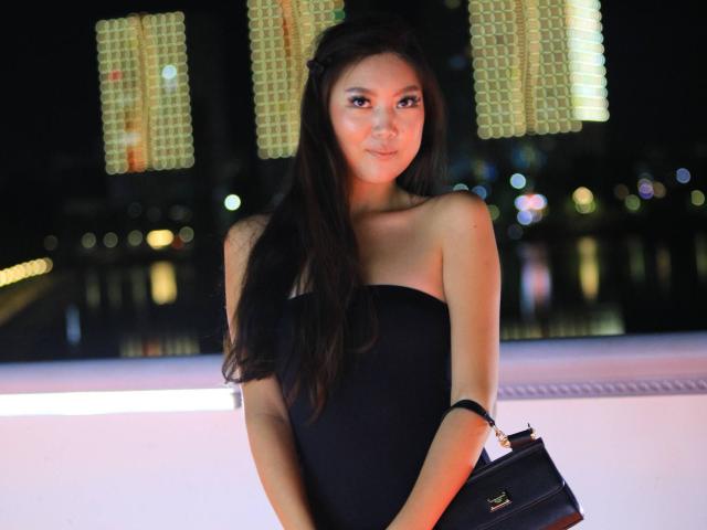 AikoTenchi with long black hair, wearing a strapless black dress, holding a black purse, smiling, night-time, city lights blurred in the background, her tits partially visible.