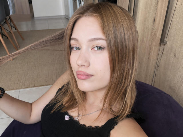 BobbyeWelson has light brown hair, green eyes, and pink lipstick. She's wearing a black lace top, holding her hair, with a bracelet on her right wrist.