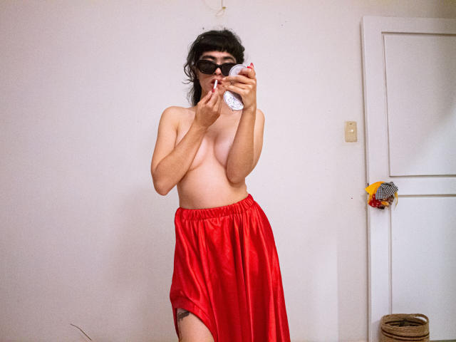 Topless FridaBiscuit met kleine borsten, donker haar, draagt een rode rok, houdt een witte mok vast, staat in een kamer, heeft een rietje in haar mond, een deur aan de rechterkant, een rieten mand rechtsonder..
