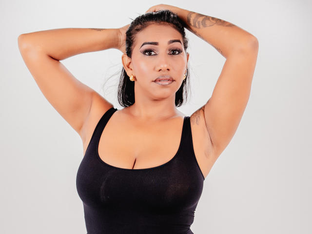 Kristelee69, les bras levés, porte un débardeur noir, dévoilant ses seins, a une peau bronzée, un tatouage sur le bras gauche, des boucles d'oreilles dorées et affiche une expression confiante. C'est une femme séduisante..