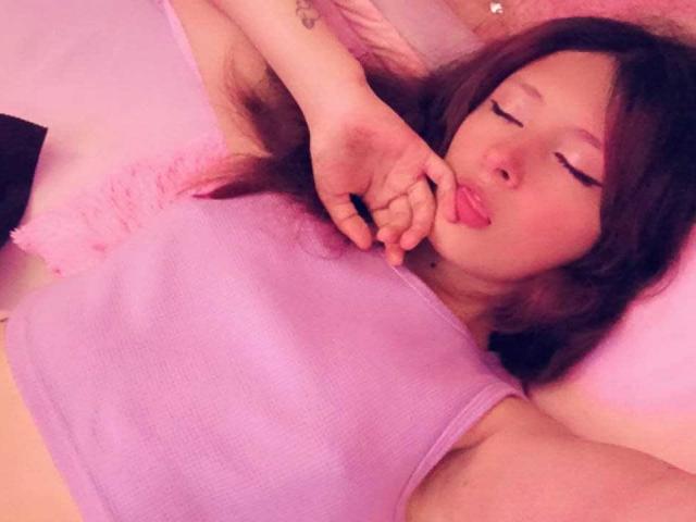 ValentinaRoseX with pinkish skin, brown hair, and closed eyes, wearing a purple tank top, pouting with tongue out, hand near chin, lying on pink pillow.
