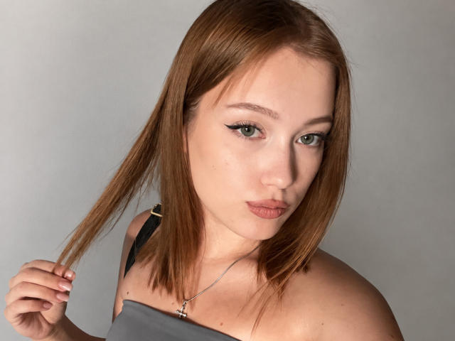 BobbyeWelson has light brown hair, green eyes, and is holding her hair with a hand. She's wearing a gray strap dress and a silver necklace.