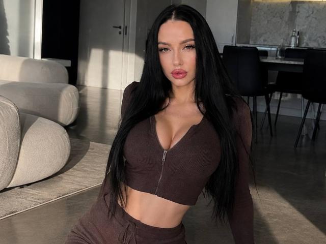 SalomeGlosson with long black hair, wearing a brown crop top and matching pants, showing her big tits and pink lipstick, sitting in a sunlit room.