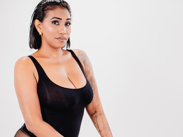 Kristelee69 tem a pele morena, cabelo preto e molhado, brincos de ouro e uma tatuagem no braço direito. Ela está vestindo uma regata preta, transparente e justa, que mostra seus seios grandes..
