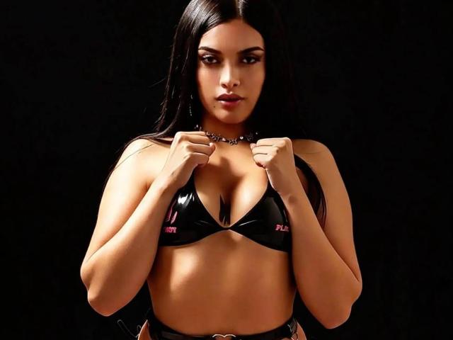 KattyRosejs, com longos cabelos pretos, usando um top de biquíni preto brilhante, segurando as alças, mostrando seus seios, olhos escuros e um colar de contas, tudo contra um fundo preto..
