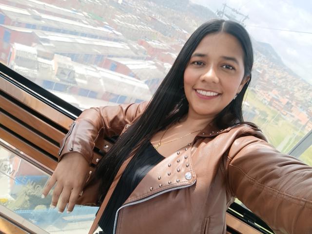 PauletteLoveX con cabello negro largo, chaqueta de cuero marrón con tachuelas, sonriendo, top negro, señalando la ventana, piel marrón media, collar de oro pequeño, busto de tamaño medio, vista exterior soleada..
