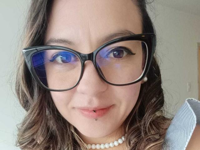 Ariana : Une blonde avec des lunettes à monture bleue, des cheveux bruns ondulés, une lèvre inférieure percée, un collier de perles et des lunettes à monture noire, regardant l'appareil photo avec un léger sourire..