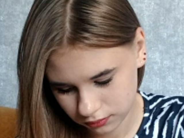 Brittaney Jecklin, con lunghi capelli castani, che guarda in basso, indossa una camicia a righe, ha il rossetto rosso e un trucco leggero, e mostra un piccolo piercing all'orecchio sinistro..