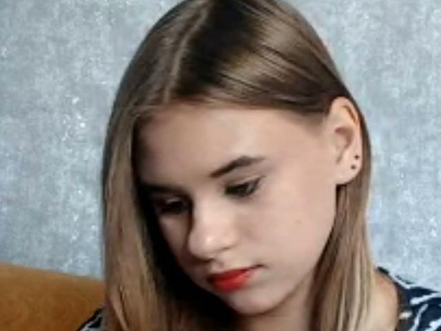 Brittaney Jecklin, com cabelo loiro, olhos baixos, batom vermelho, pequenas argolas nos ouvidos e uma pequena perfuração no nariz, vestindo uma camisa azul com estampas, olhando para baixo..