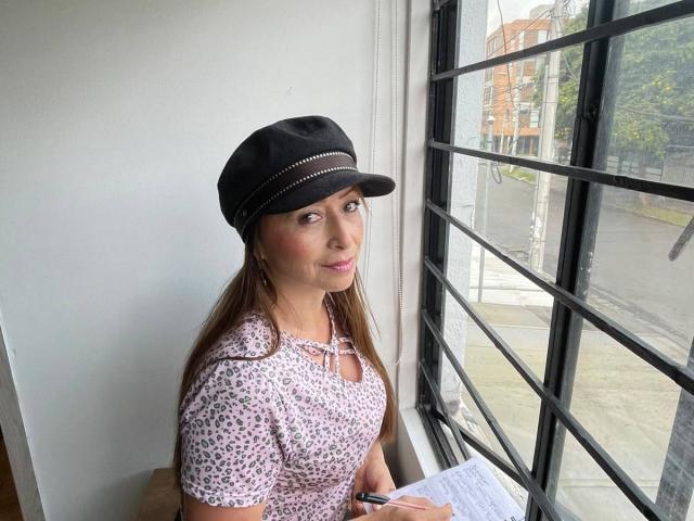 GianineLarose with brown hair, wearing a black cap and pink leopard print top, sitting by a window, holding a pen and paper, looking slightly to the side.