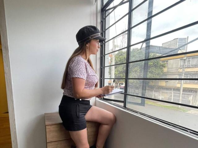 GianineLarose sits by a window, wearing a black cap, pink patterned top, black shorts, holding a notebook, with long brown hair, looking outside. Her ass is visible.