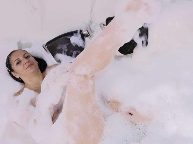 MadameScarlett is naked in a bubble-filled bathtub, legs up, exposing her pussy. She has dark hair, a light skin tone, and a relaxed, slightly smiling expression.
