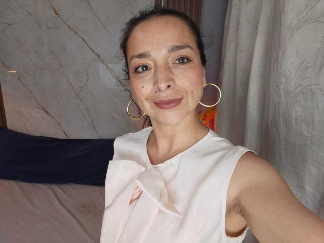 StangelarRivera with dark hair, hoop earrings, white sleeveless top, slight smile, visible armpit hair, light skin, brown eyes, small nose, pink lipstick, subtle freckles.