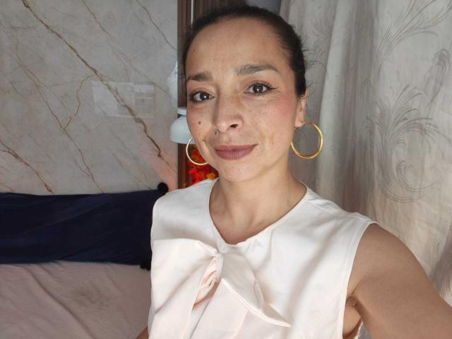 StangelarRivera has brown skin, freckles, and small tits, wearing a white sleeveless shirt with a pink bow, gold hoop earrings, and a subtle smile in a bedroom.