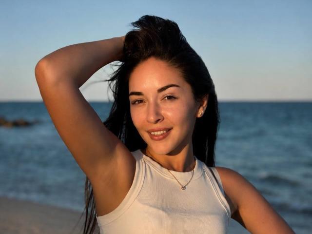 AquaLady with tan skin, black hair, and a smile, wearing a white tank top, raising her right arm, showing her armpit, and a small necklace against a blue ocean.