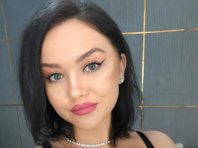 JessyQuinn tem cabelo preto, olhos azuis e lábios rosados. Ela está usando um colar de pérolas e brincos de diamantes, com delineador marcante e um sorriso discreto. Seus seios não estão visíveis..