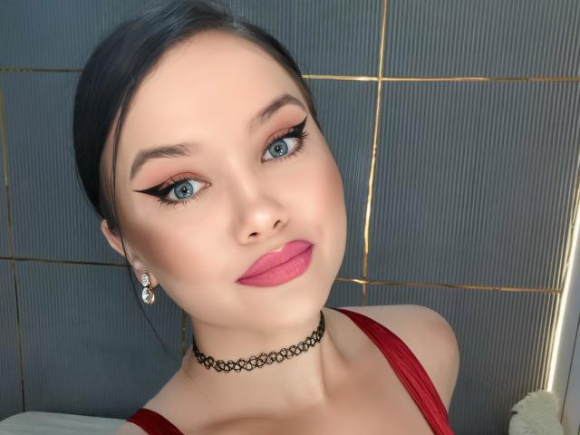 JessyQuinn, con gli occhi azzurri, il rossetto rosa, la collana nera, il vestito rosso e gli orecchini di diamanti, mostra il suo seno, ha i capelli neri e un viso sexy, valorizzato dal trucco..