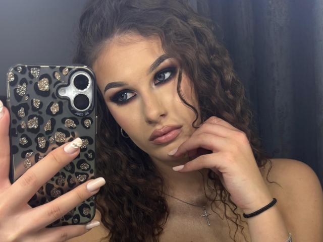 CharlotteeW, con cabello castaño rizado, sosteniendo un teléfono con una funda de estampado de leopardo, mirando en el espejo, con maquillaje de ojos azul, lápiz labial rosa, un collar con una cruz y una goma para el pelo negra en la muñeca..
