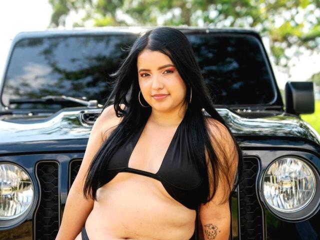 JuicyCurvaceous, eine mollige lateinamerikanische Frau mit langen schwarzen Haaren, trägt einen schwarzen Bikini-Oberteil und steht vor einem schwarzen Jeep, wobei sie ihre kleinen Brüste und ihren Bauch zeigt..