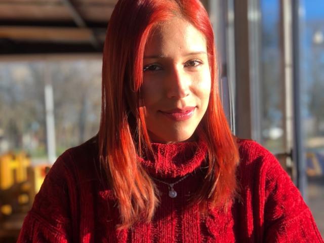 KiaraHernandezX with red hair, smiling, wearing a red knit turtleneck, sunlight highlighting her face, necklace with small pendant, background blurred, casual indoor setting.
