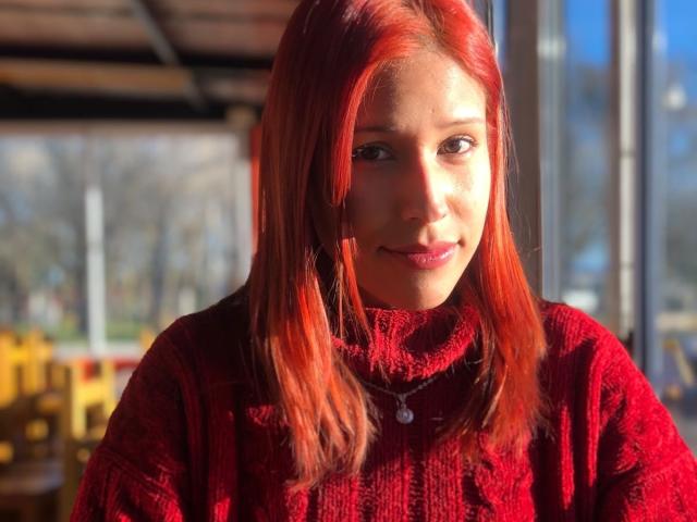 KiaraHernandezX with red hair, wearing a red turtleneck, has a cute smile, and a silver necklace, sitting by a window, sunlight highlighting her face.