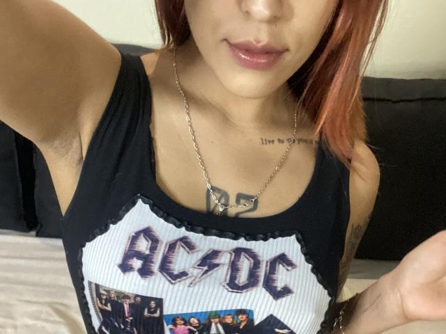 KiaraHernandezX with red hair, black ACSO tank top, shiny pink lips, silver necklace, armpit hair, small tattoo on chest, bed in background. Boobs, chest, tits visible.