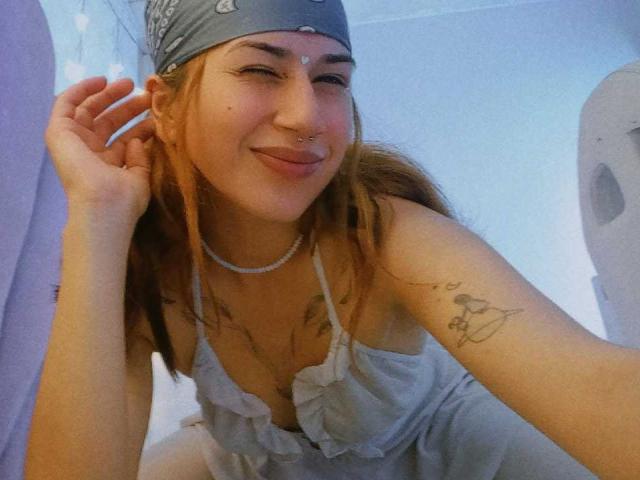 NaraStorn, com um piercing no nariz, maquiagem nos olhos e um lenço azul na cabeça, sorrindo, vestindo uma blusa branca com babados, mostrando uma tatuagem no braço e um colar de pérolas..