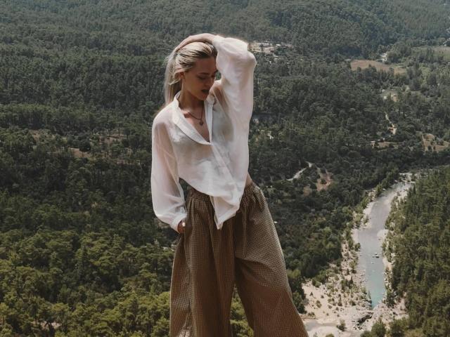ThomasinaOrtz with blonde hair, white loose blouse, and brown high-waisted pants, hand in hair, standing, looking down, forested mountain background, river visible.