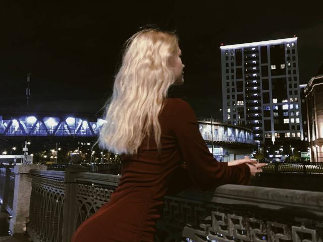 ThomasinaOrtz with long, wavy blonde hair, wearing a tight, dark red dress, leaning on a bridge railing, facing away, showing her ass, at night, city lights in background.