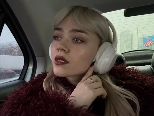 ThomasinaOrtz with white headphones, blonde hair, red lipstick, and a fluffy red sweater, looking out the car window with a slight smile, holding her earpiece.