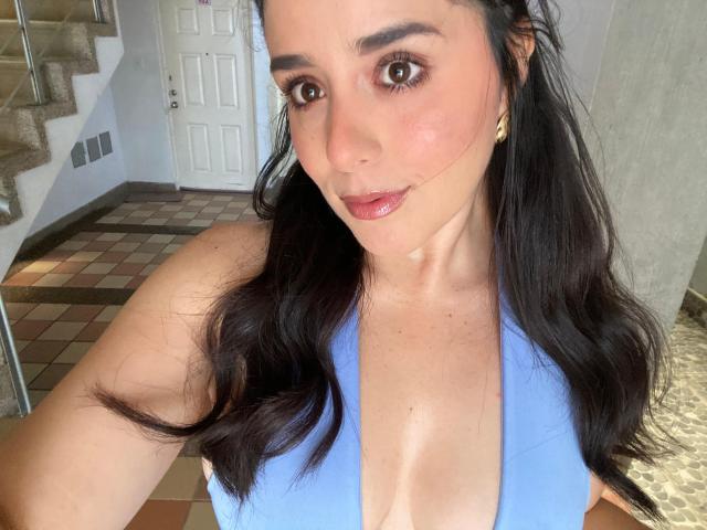 VickyMegan with dark hair, blue top, and cleavage showing her shiny, plump tits in a selfie. She's got a cute face, brown eyes, and glossy pink lips.