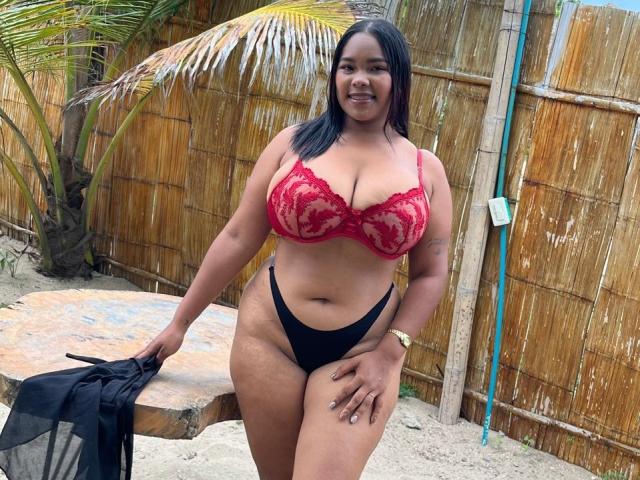 LaikaMar, eine kräftige, schwarze Frau mit großen Brüsten, trägt einen roten Spitzen-BH und einen schwarzen String. Sie hält ein schwarzes Netzoberteil in der Hand und steht auf sandigem Boden. Im Hintergrund ist ein Bambuszaun zu sehen. Sie lächelt..