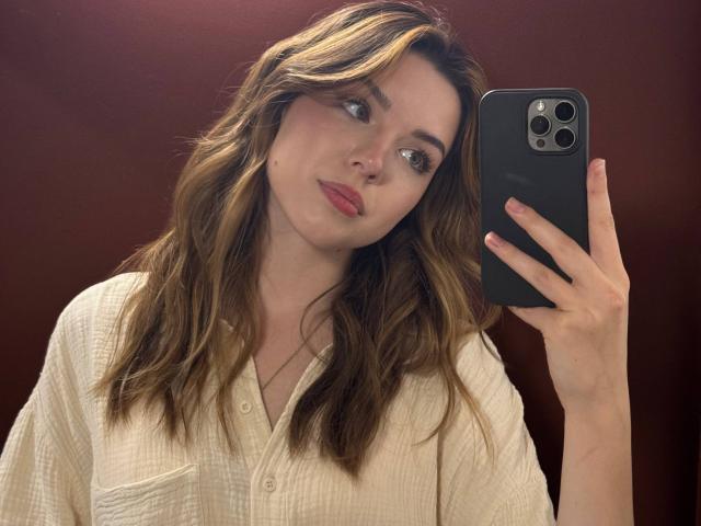 EviaDorland, met golvend bruin haar, roze lippen en een wit overhemd, houdt een zwarte telefoon vast en maakt een selfie, waarbij ze haar borsten in de spiegel laat zien..