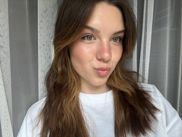 EviaDorland, met volle lippen, bruin golvend haar, groene ogen en een witte blouse, met een klein beauty markje op haar rechterwang. Ze maakt een lichtjes fronsende grimas..