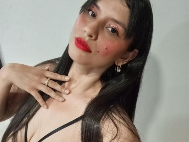 MadissonFontaine with long black hair, red lipstick, and pink blush, wearing a black strap top, hand on chest, gold earrings, and a gold ring, looking at camera.