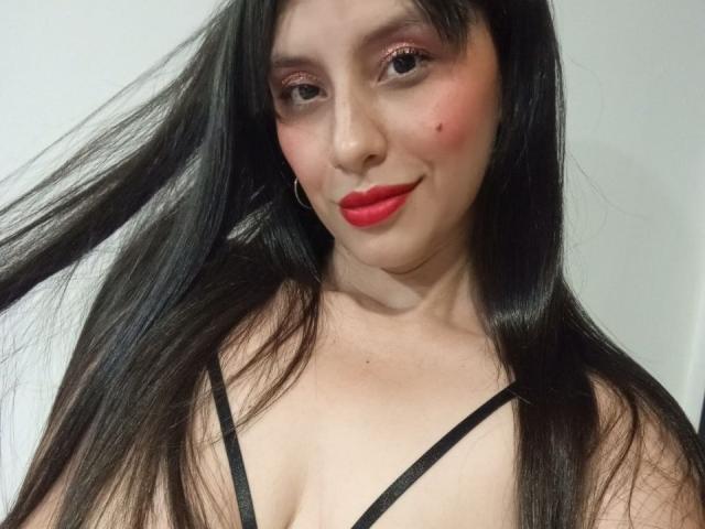 MadissonFontaine with long black hair, red lipstick, pink eye shadow, small black mole on her right cheek, black strappy top, smiling, holding hair on the left.