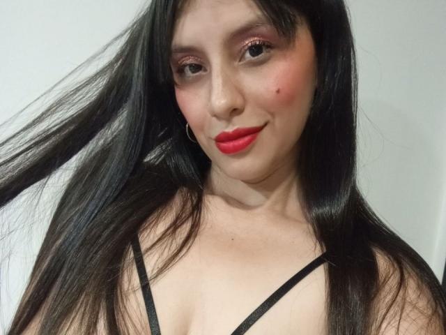 MadissonFontaine with long black hair, red lipstick, pink eyeshadow, small mole on cheek, smiling, black strappy top, holding hair, gold hoop earring, light skin.