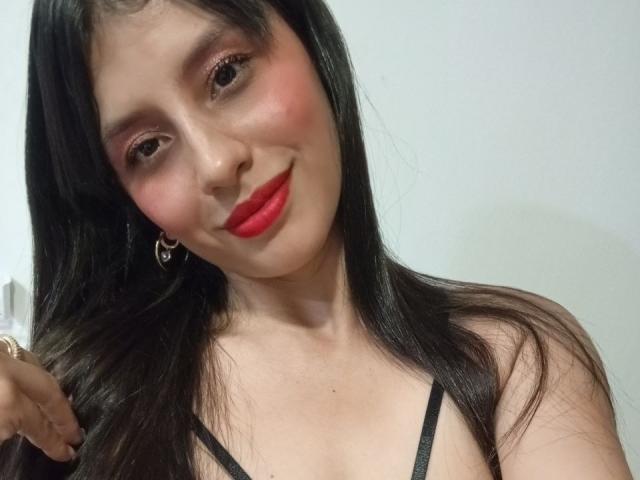 MadissonFontaine with long black hair, red lipstick, pink eyeshadow, and a small smile, wearing a black spaghetti strap top, holding her hair with a gold earring visible.