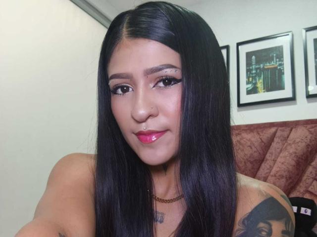 LuciadelaVega with long black hair, pink lipstick, black eyeliner, nose ring, small tattoo on right shoulder, and gold necklace, smiling softly at the camera.