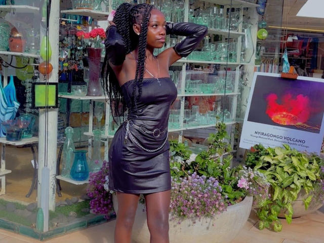 EbonyAngel-hot está parada en una tienda, vistiendo un vestido ajustado, negro, con un hombro descubierto, de cuero, con mangas largas y detalles de cadena. Tiene el pelo largo, trenzado, y una pose segura..