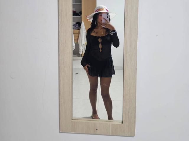 MyaaTyler met donkere huid, een zwarte doorschijnende top met uitsparingen, een zwarte rok, blote benen, een zonnehoed, die een telefoon vasthoudt en in de spiegel kijkt, staat zonder schoenen en kijkt naar haar reflectie..