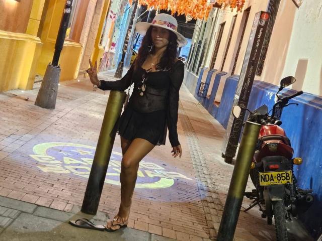 MyaaTyler standing on a cobblestone street, wearing a black sheer top, black skirt, sun hat, and flip flops, showing a peace sign, with a red motorcycle beside her.