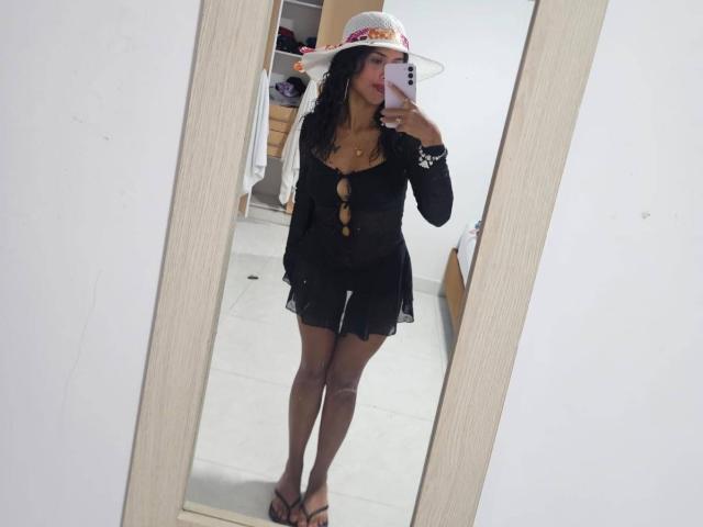 MyaaTyler with black sheer dress, cleft front, showing tits, wearing a white sunhat with flowers, black stockings, and sandals, standing in front of a mirror.