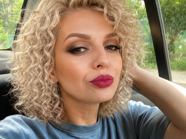 DouceAmelia ha i capelli biondi ricci, l'eyeliner scuro e il rossetto rosa. Indossa una camicia blu e guarda la fotocamera con un leggero sorriso. Ha una mano dietro la testa..